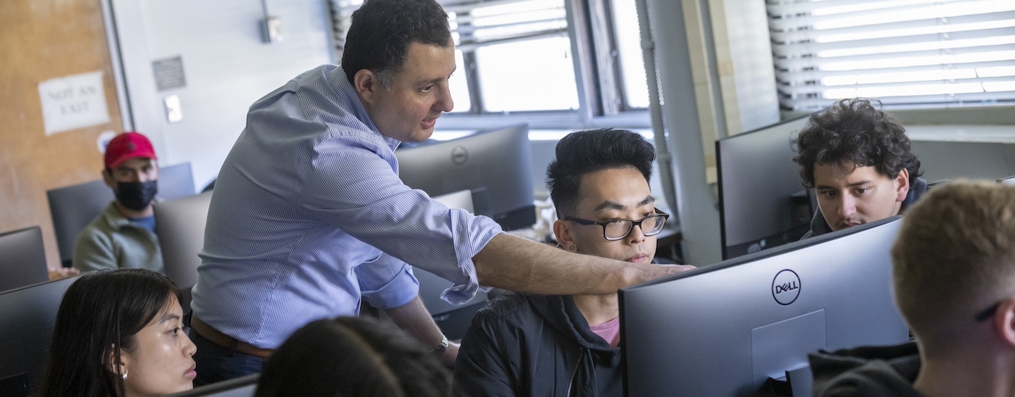 students in a classroom at computers, with instructor in the middle of the room, pointing at a screen