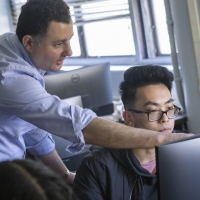 students in a classroom at computers, with instructor in the middle of the room, pointing at a screen