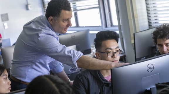 students in a classroom at computers, with instructor in the middle of the room, pointing at a screen